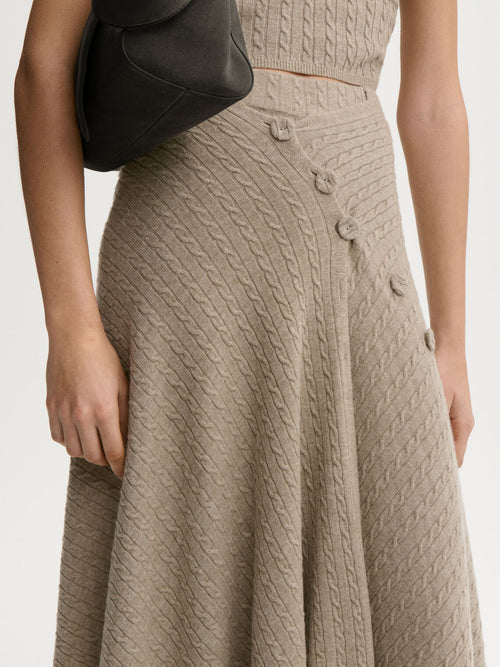 LIZA sjal/cape fra By Malene Birger i fargen beige, laget i 90% ull og 10% cashmere. Kabelstrikket og lukkes med trukne knapper – kan brukes som sjal over skuldrene eller som skjørt rundt livet.
