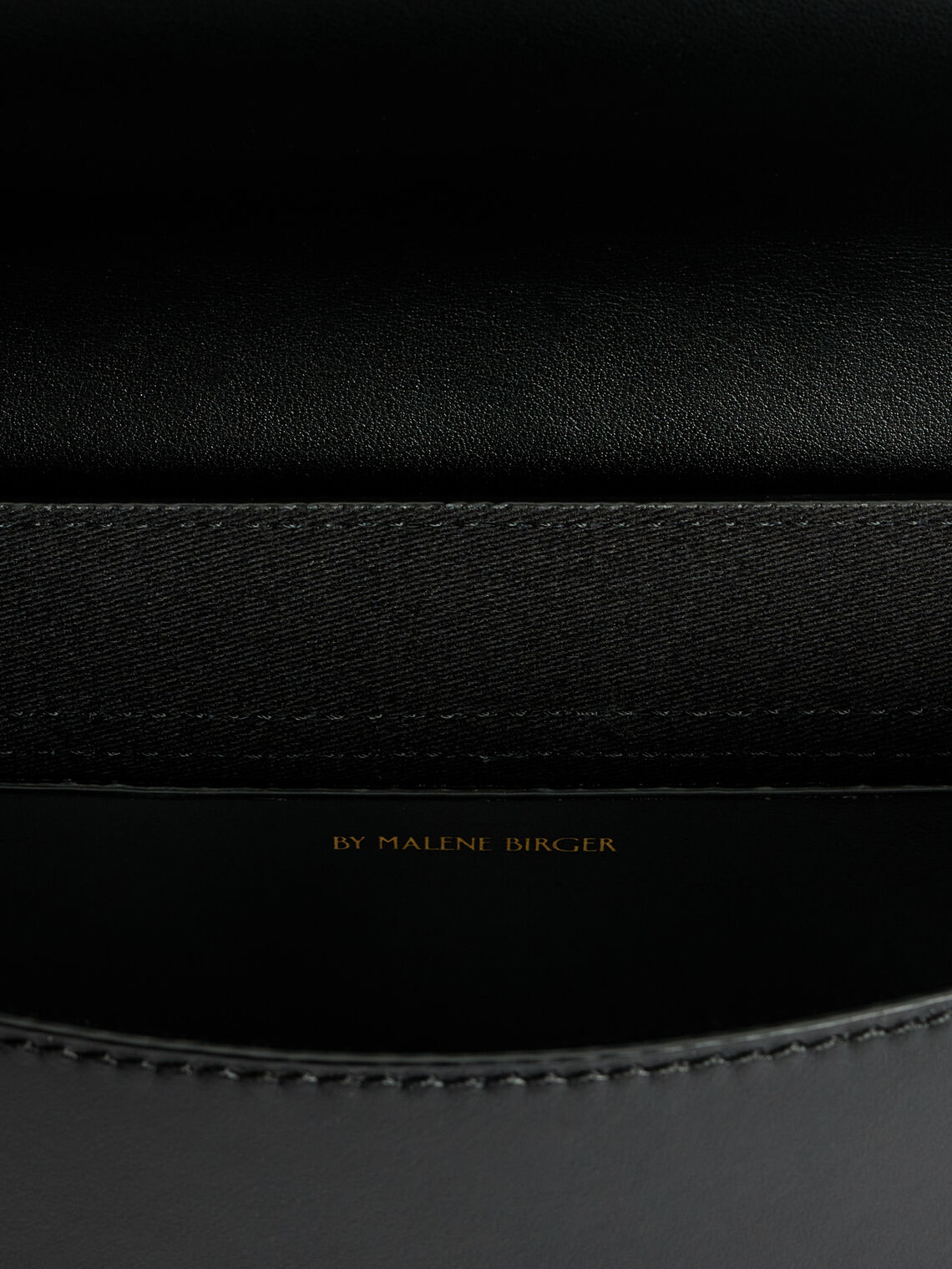 By Malene Birger Maellon veske i blankt, sort kuskinn, med diskret monogram i front og justerbar stropp.

