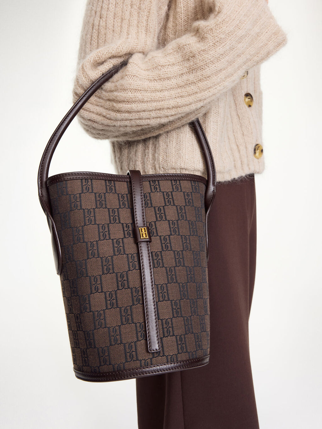 By Malene Birger Marylle bøtteveske i brun, laget av resirkulert bomull og polyester, med et gullfarget monogram-emblem i front.
