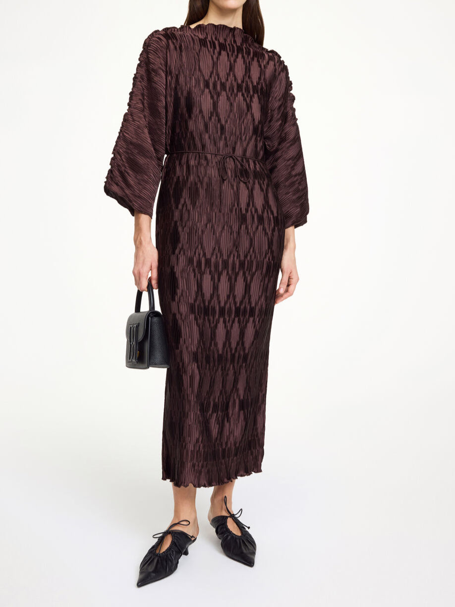 By Malene Birger Tralaido kjole i mørkebrun, med plissé struktur, båtutringning og vide ermer, laget av resirkulert polyester.
