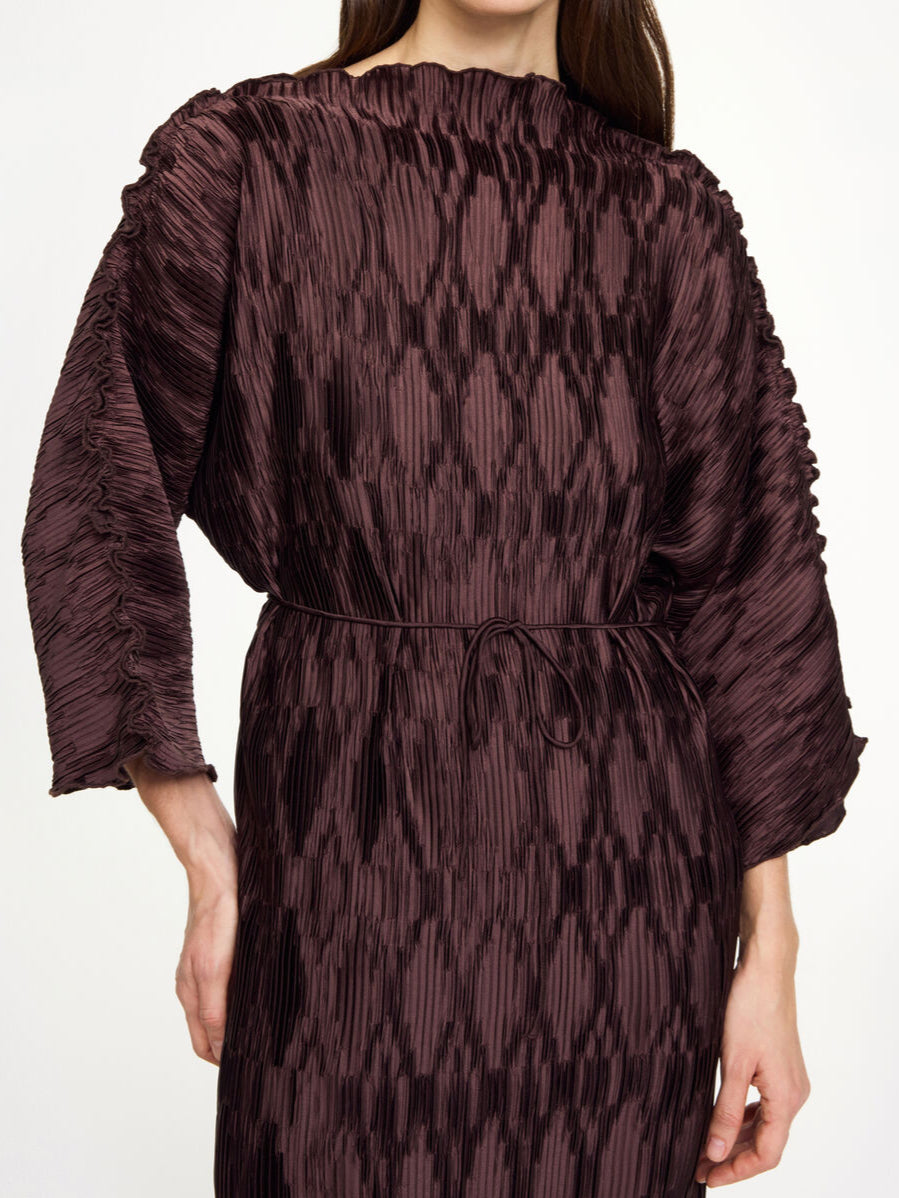 By Malene Birger Tralaido kjole i mørkebrun, med plissé struktur, båtutringning og vide ermer, laget av resirkulert polyester.